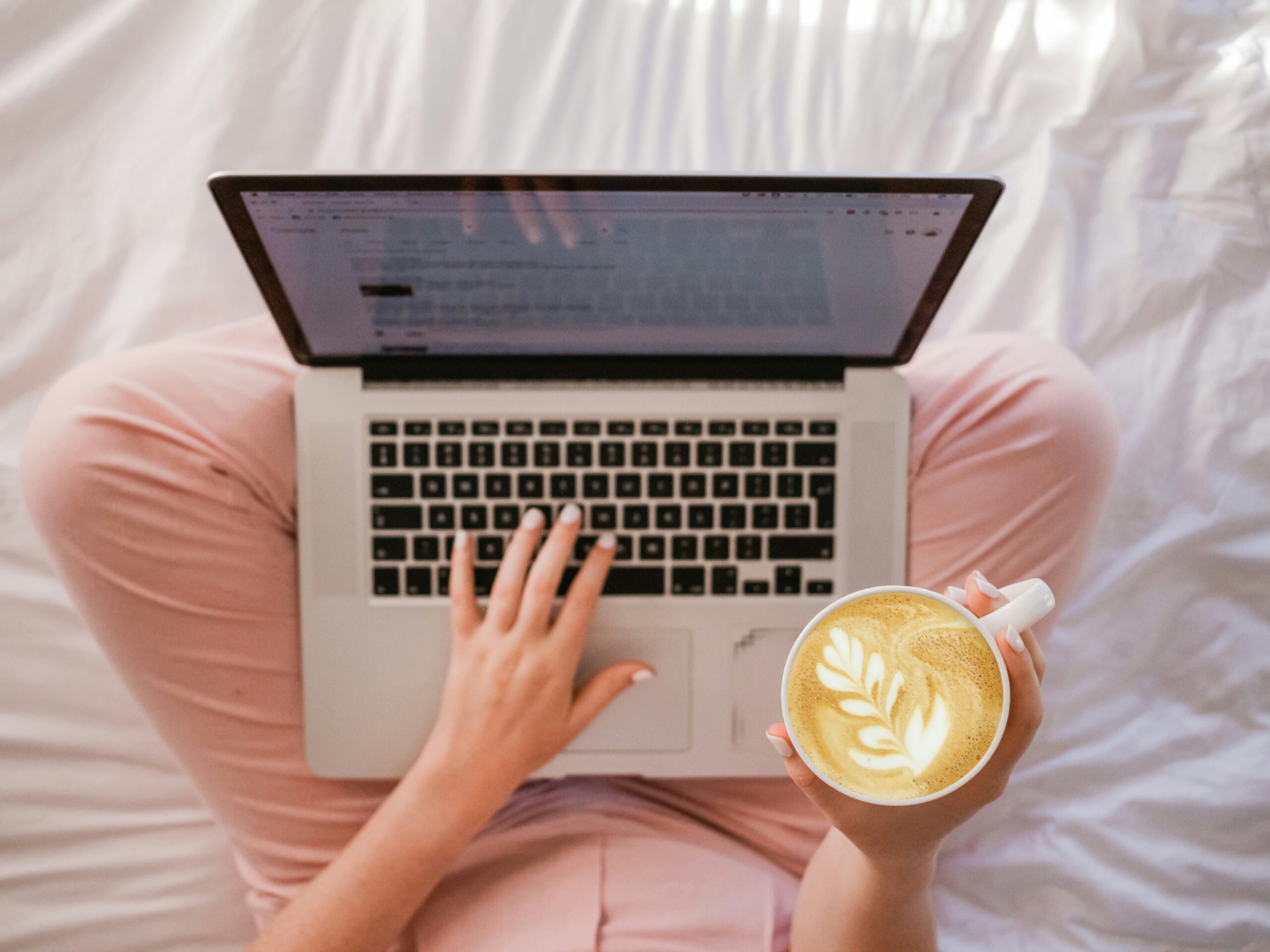A photo of an aerial view of a person sitting on a bed with their legs crossed, with a laptop on their leg, holding a cup of coffee.